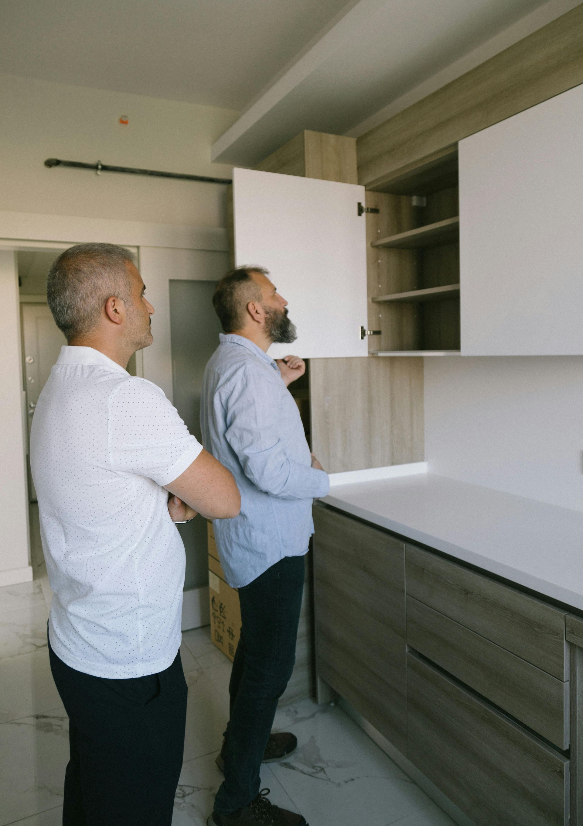 Two men examining a kitchen cabinet. One opens it; the other observes. Light wood and white cabinets.