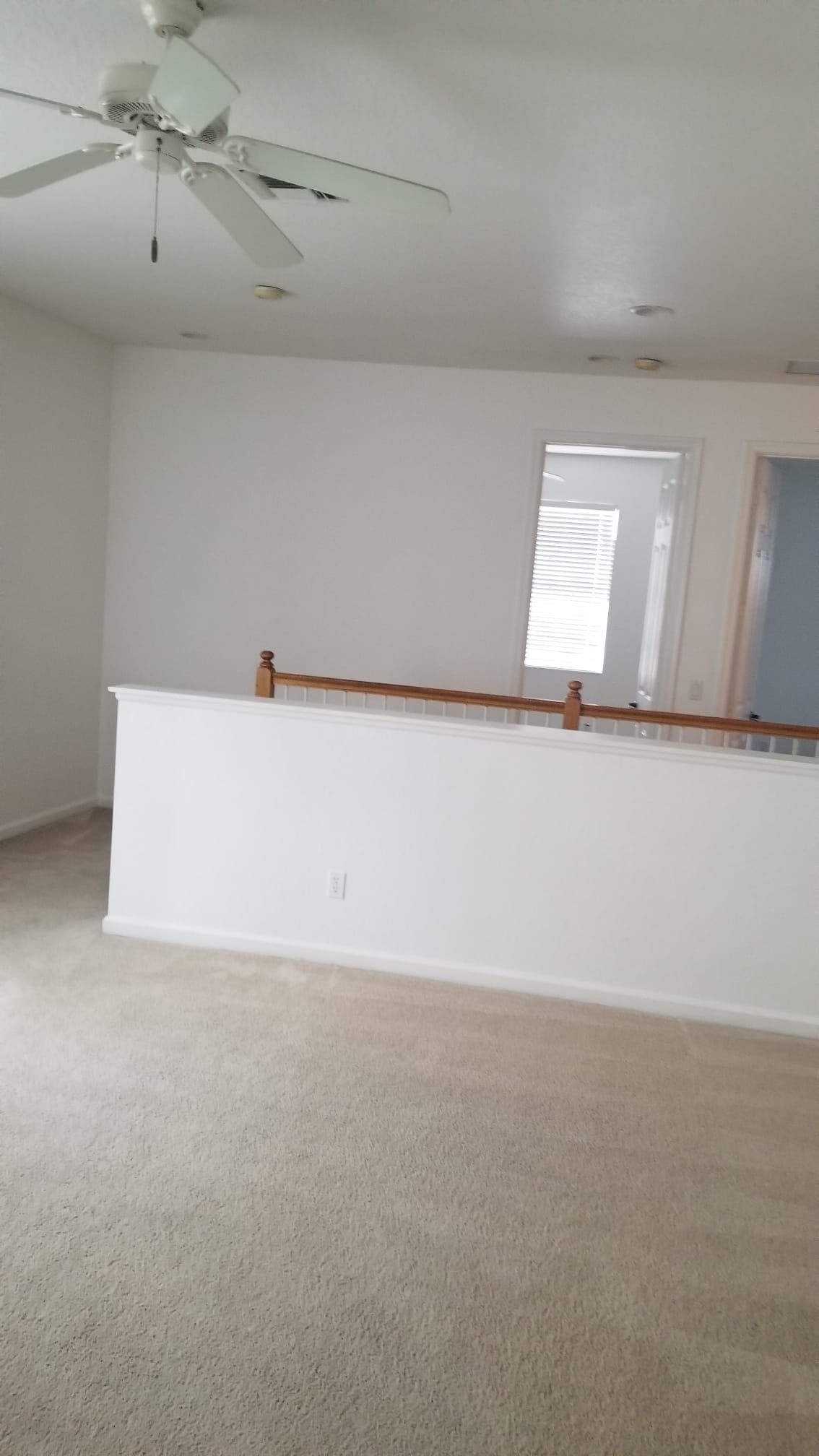 Empty living room with white walls, carpet, and a half wall with wooden railing. A ceiling fan is visible.