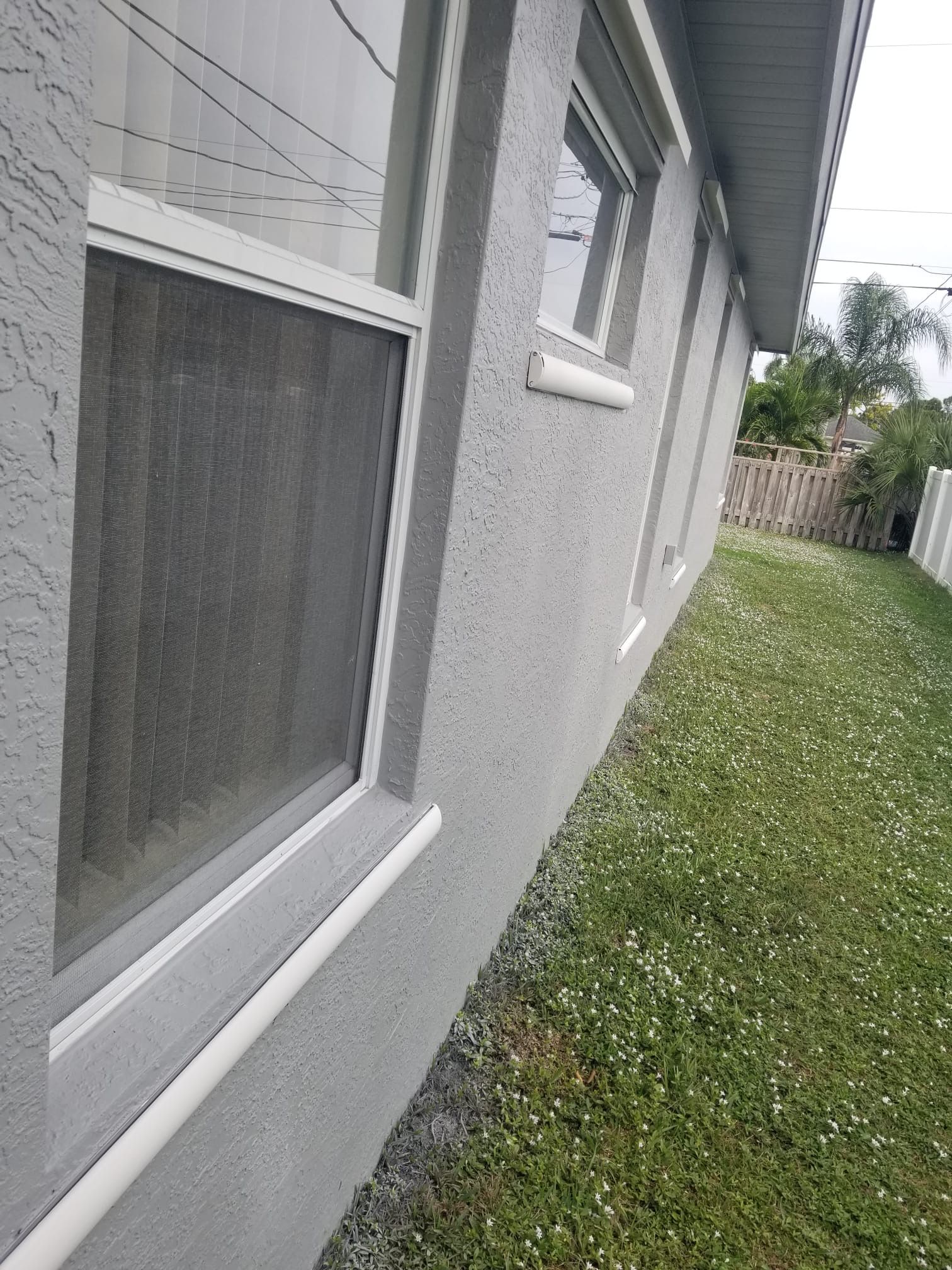 Gray stucco house exterior with windows, next to a grassy lawn with white flowers and a fence.