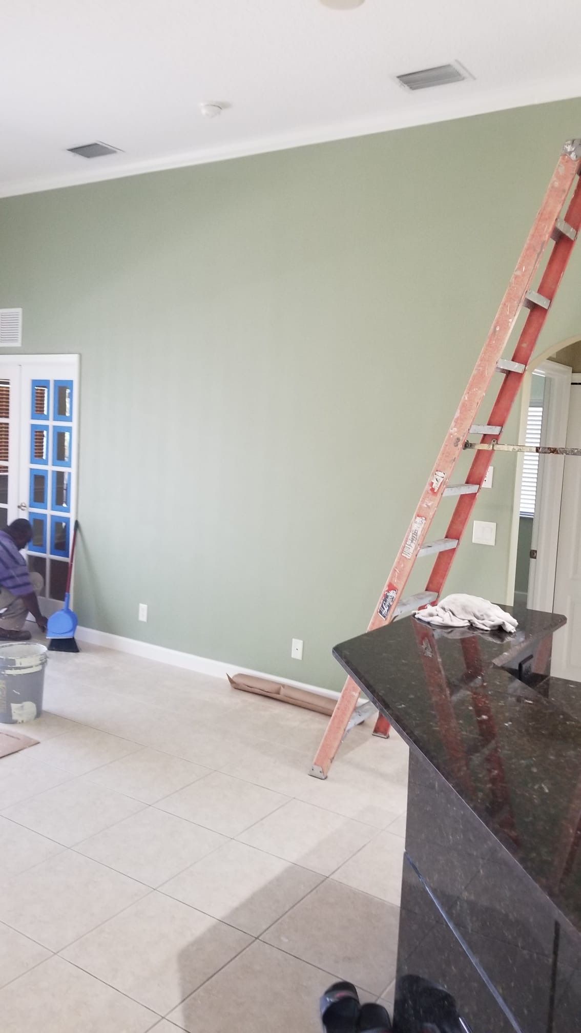 A person paints a green wall; a ladder leans against it.  A counter and door are also visible.