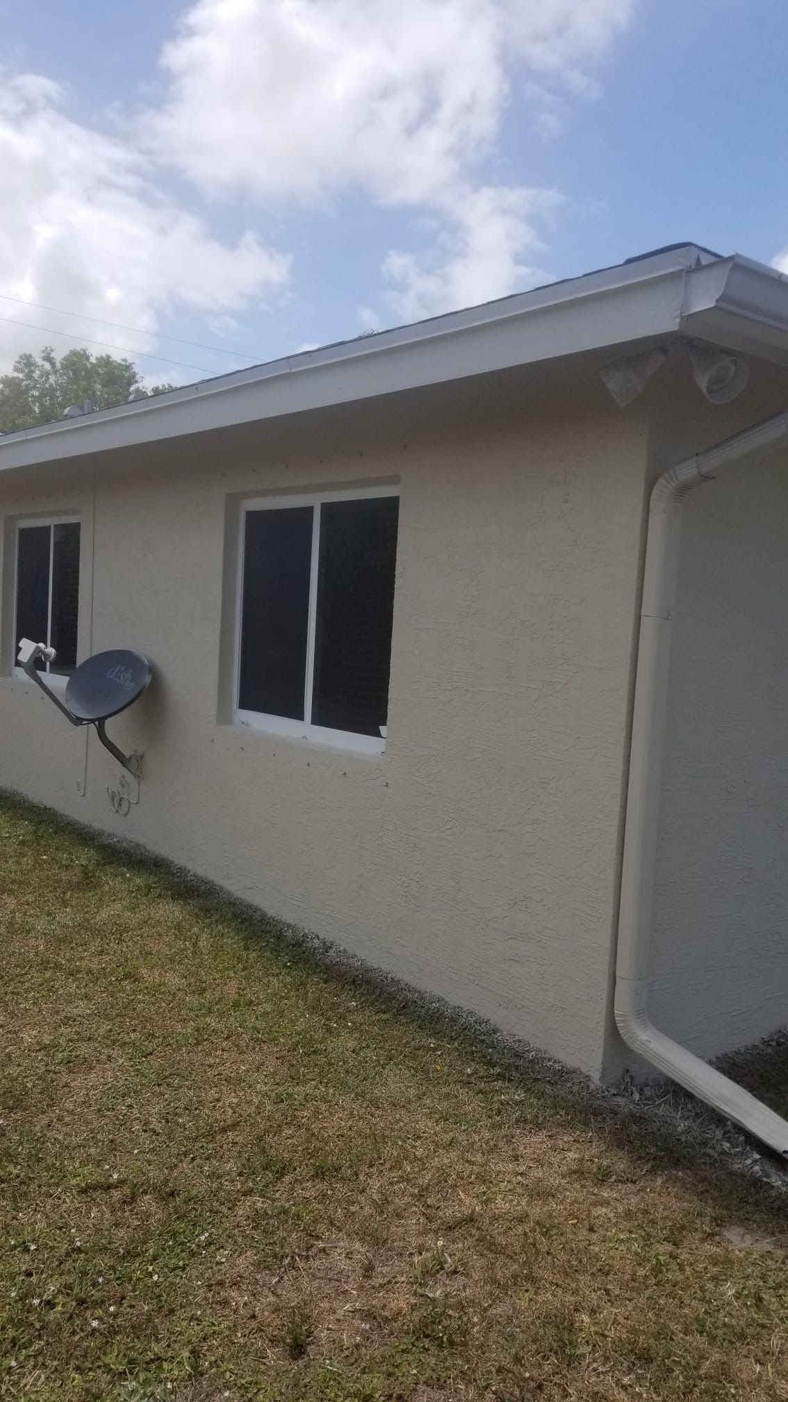 Tan stucco building with two windows and a satellite dish on a grassy lawn under a partly cloudy sky.
