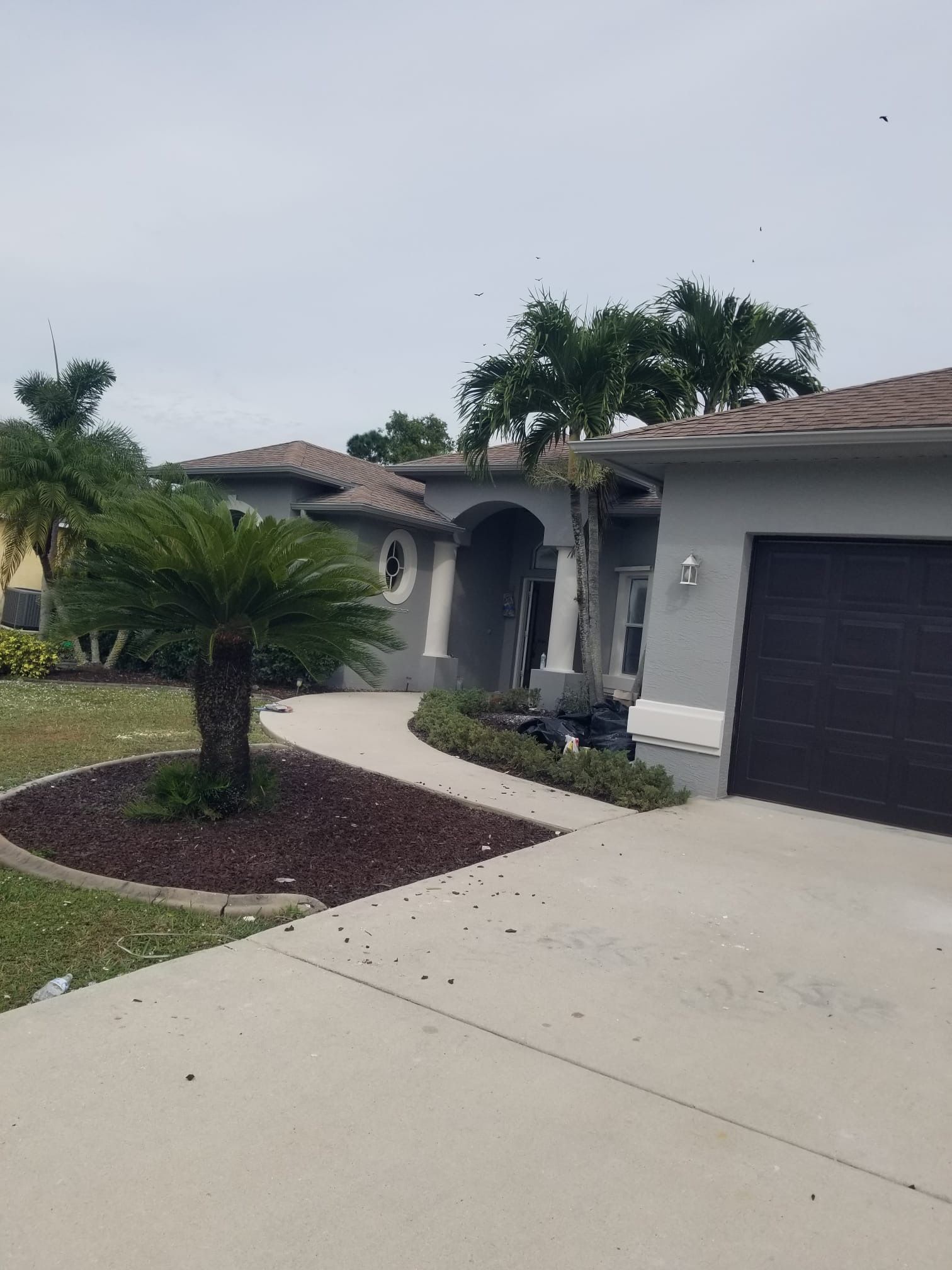 Gray house with brown roof, driveway, and landscaping. Palm trees and overcast sky.