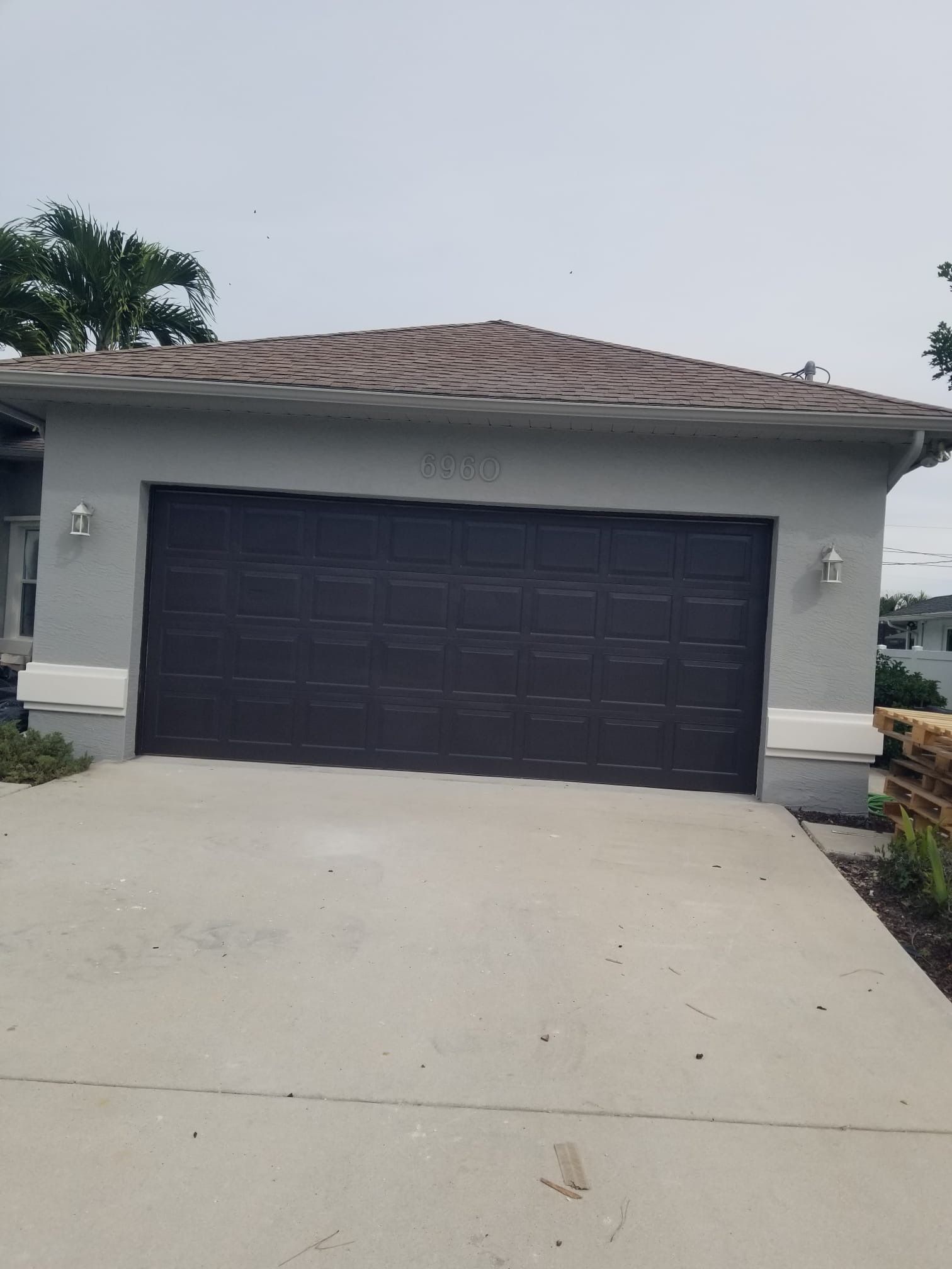 A light gray house with a brown garage door on a concrete driveway, under a cloudy sky.