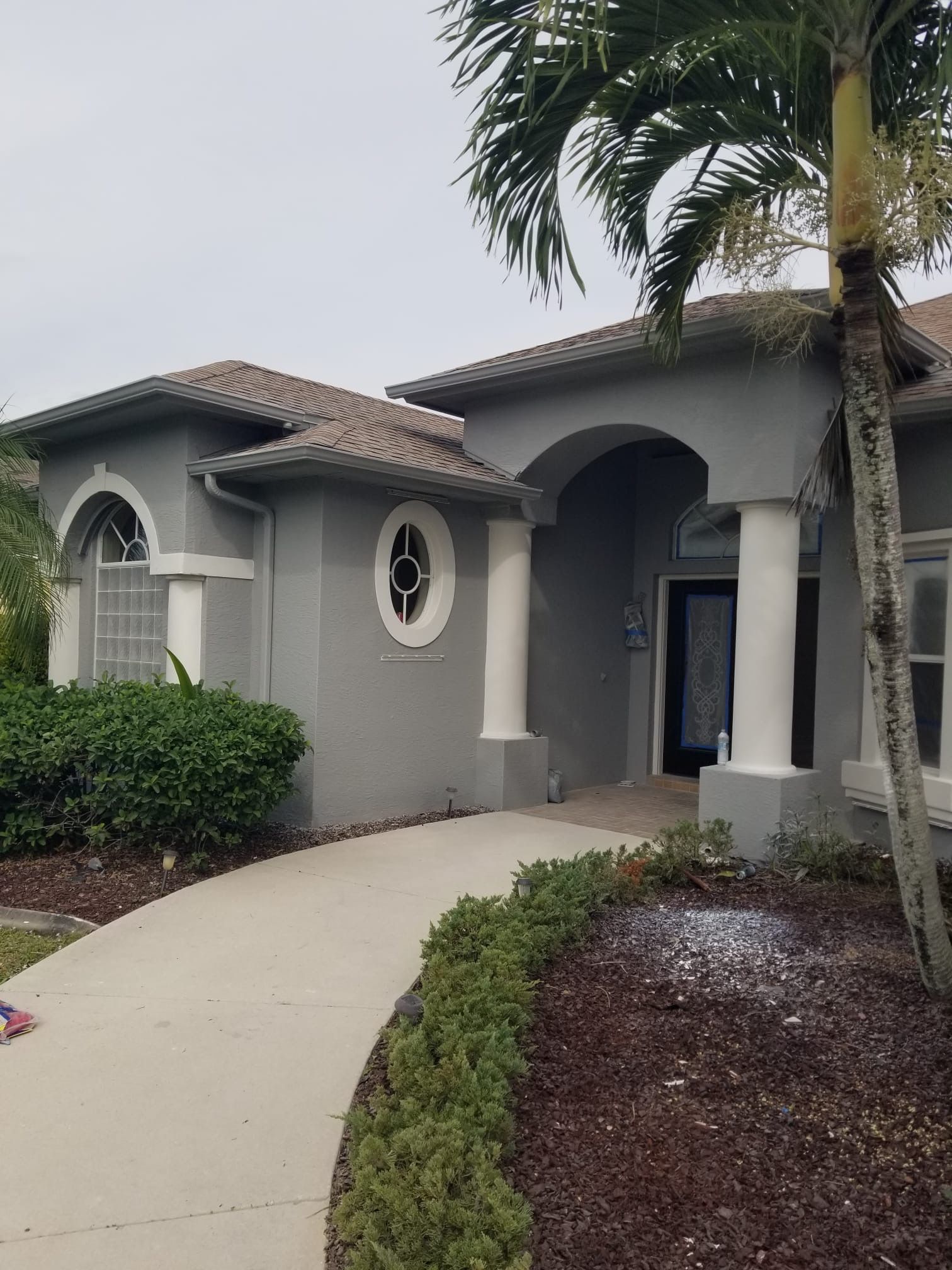 Gray stucco house with arched doorway, white columns, and curved walkway lined with greenery. Palm tree on the right.