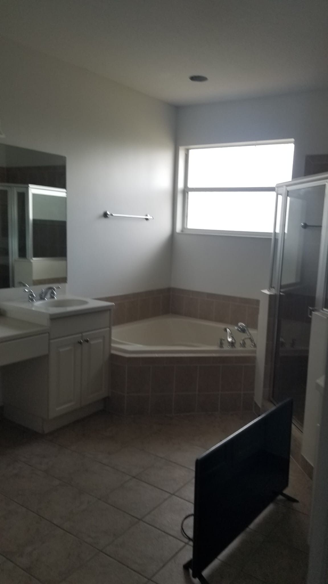 Bathroom with bathtub, shower, vanity, and TV. Tan tile floor, light walls, and a window.