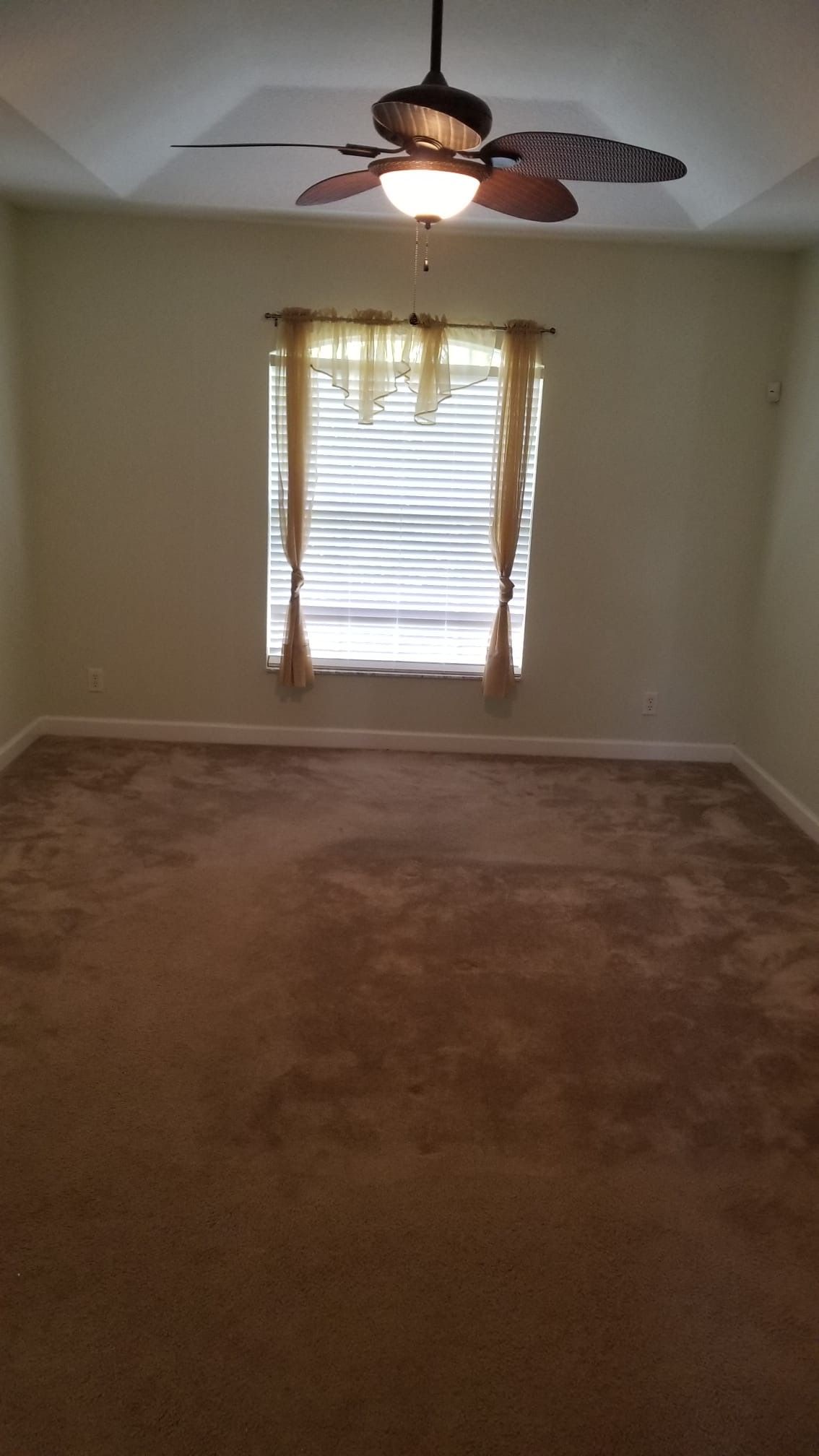 Empty bedroom with brown carpet, window with blinds and curtains, and ceiling fan.